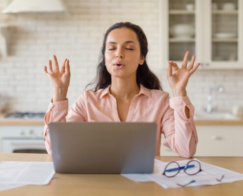 Woman practicing deep breathing meditation while sitting at her laptop in a kitchen. A women's therapist in Medford, NJ can help women struggling with parental burnout and more. Get started today with a supportive therapist. 