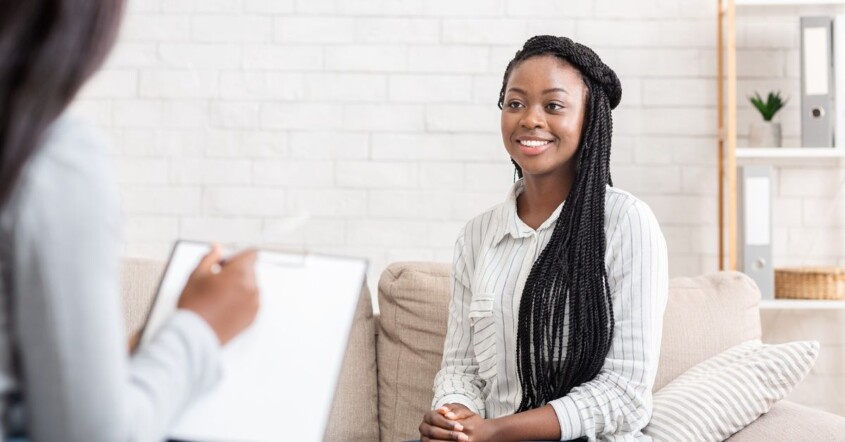 A woman sits on a sofa smiling as she speaks with a therapist during a counseling session