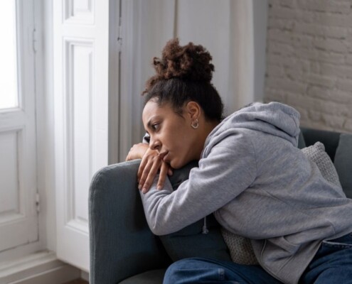 A woman experiencing sadness and emotional stress while sitting alone and leaning on the armrest of a couch at home