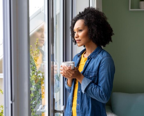 A woman stands by a window holding a mug, looking outside with a calm, reflective expression