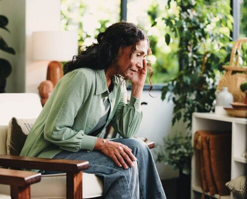 Older woman sitting alone at home looking stressed with eyes closed while holding her face, illustrating emotional burden and long-term mental load