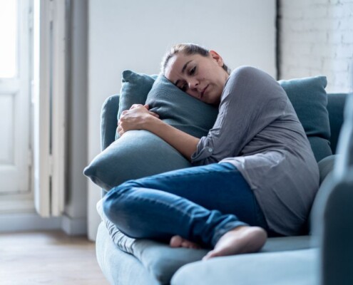 Woman lying on couch hugging pillow looking emotionally drained, representing mental load, stress, and burnout at home