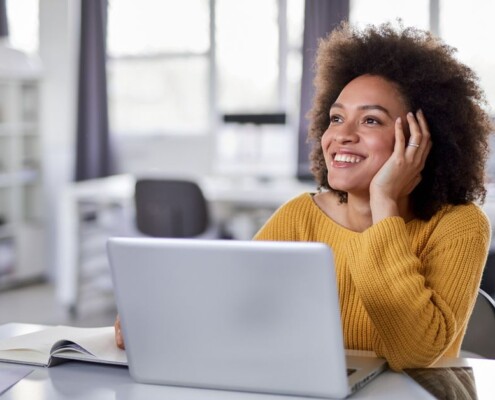 Woman sitting in front on laptop in office looking thoughtful and distracted, staring out into space while smiling