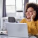 Woman sitting in front on laptop in office looking thoughtful and distracted, staring out into space while smiling