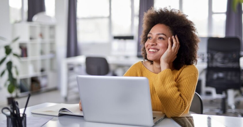 Woman sitting in front on laptop in office looking thoughtful and distracted, staring out into space while smiling