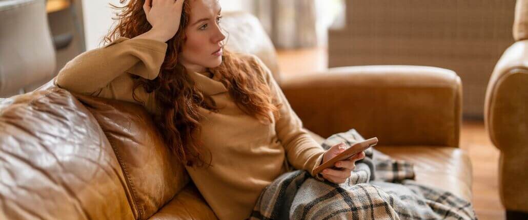Woman sitting on couch looking stressed while holding phone, representing mental load, overthinking, and emotional strain at home