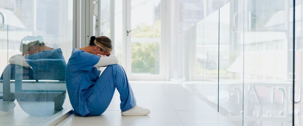 Exhausted healthcare worker sitting alone in a hallway with head down, illustrating burnout, emotional strain, and mental load in healthcare.