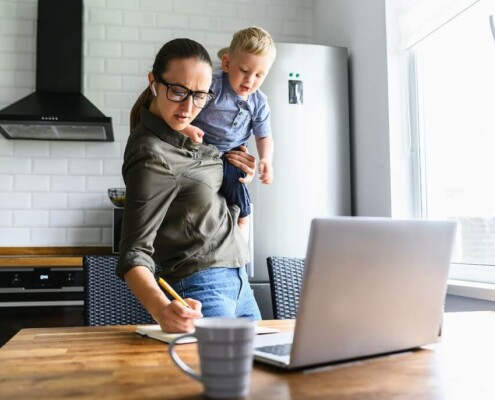 Working mom holding baby on hip while using laptop at home, illustrating mental load, multitasking, and caregiving stress