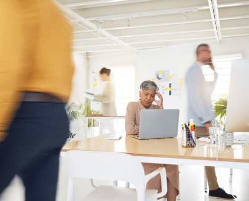 Overwhelmed older professional woman sitting at a desk in a busy office, experiencing workplace stress