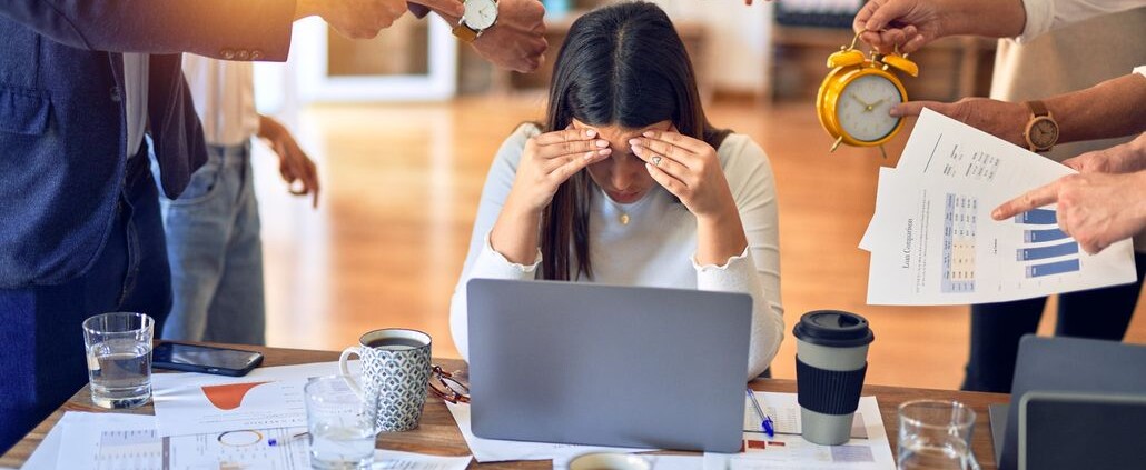 Stressed woman at laptop surrounded by coworkers demanding attention with deadlines, paperwork & more. Work burnout can affect women's relationships & home life. A therapist for women in Medford, NJ can support you today in navigating stress & burnout. 